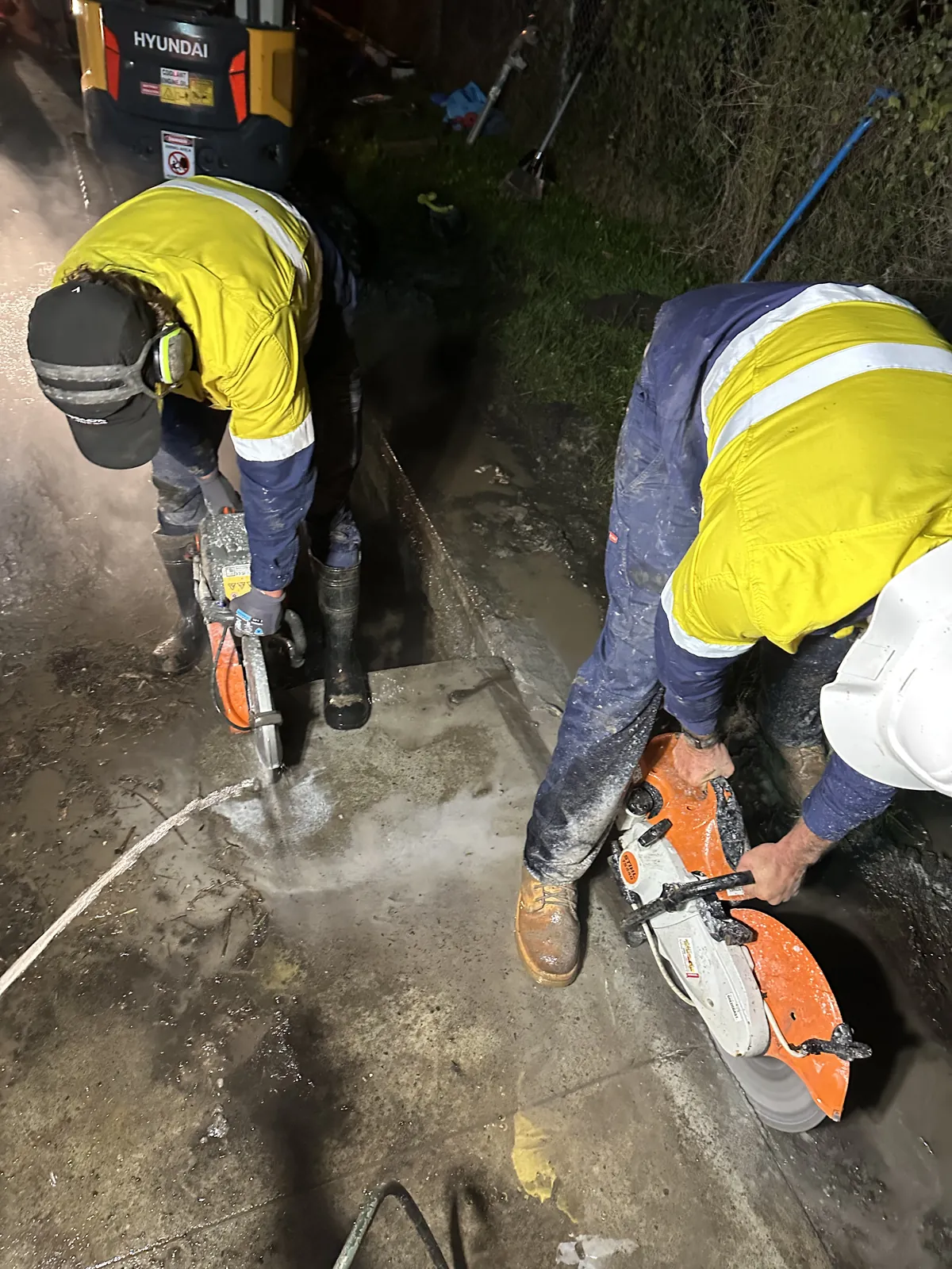 Two workers in high-visibility clothing using orange concrete saws on a concrete surface at night