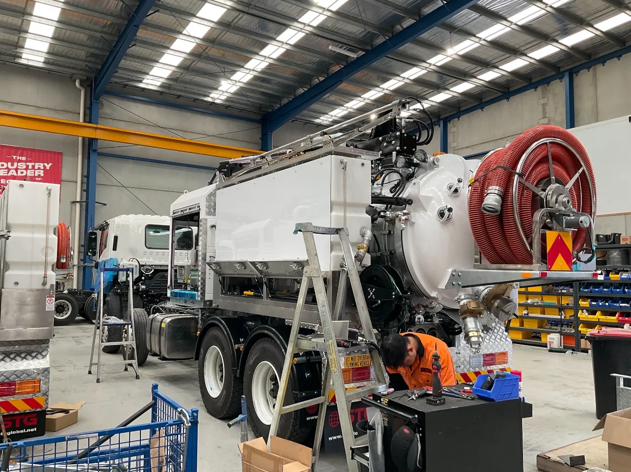 Vacuum tank fit-out on the hydro vac truck in the workshop Rear three-quarter view of the hydro vac truck with its vacuum tank, red hose reel and a worker beside a tool trolley