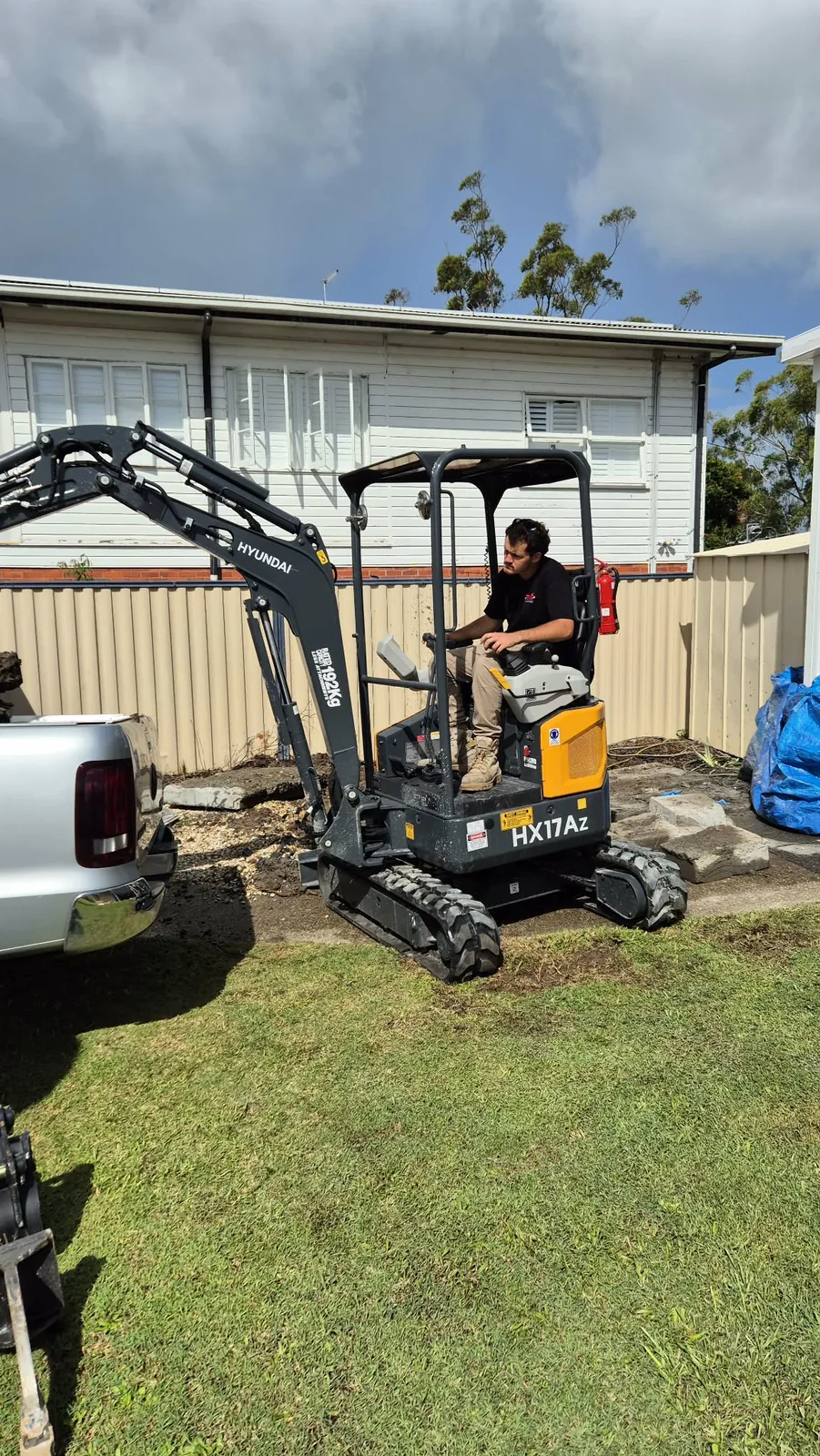 Operator seated in a Hyundai mini excavator on grass beside a ute and garage in a residential yard