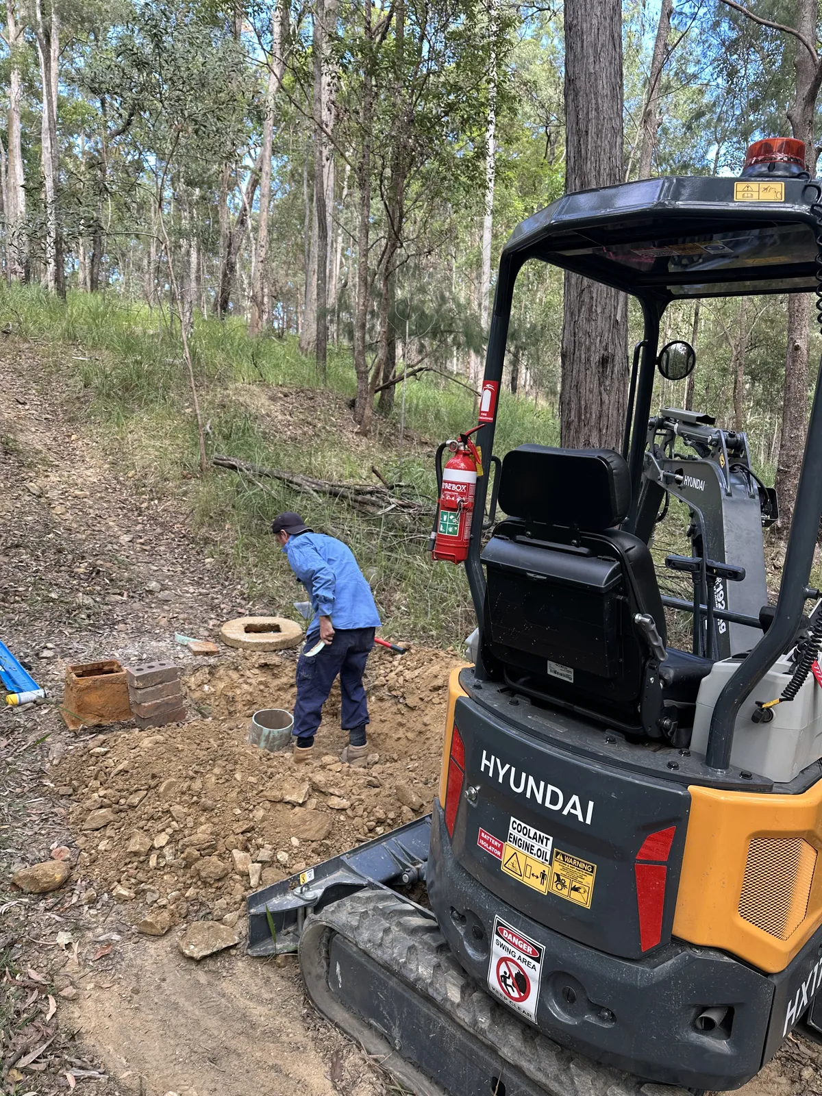 Mini excavator beside bush-track service-pit work Hyundai mini excavator beside a worker and exposed service pits along a wooded track