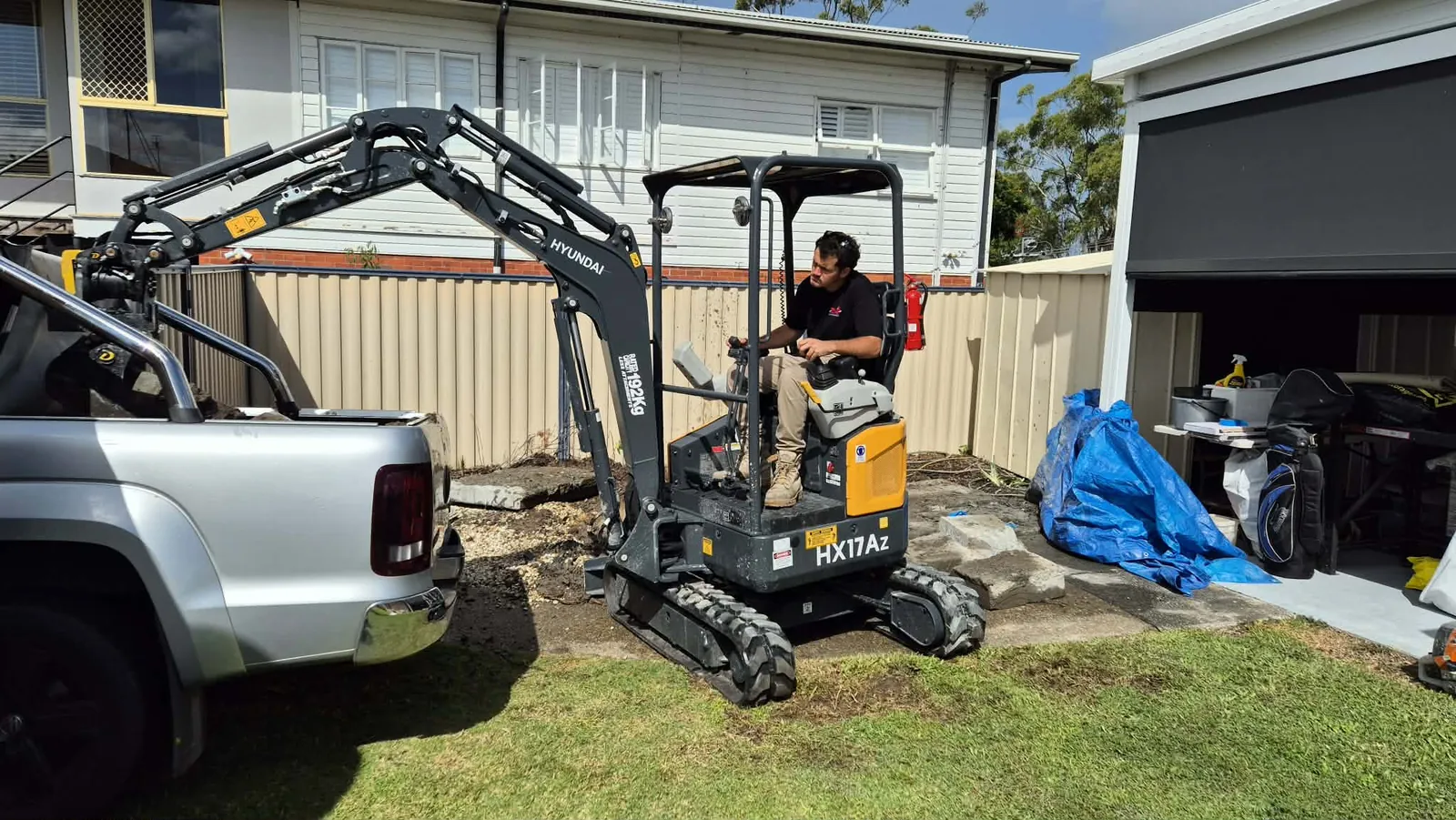 Mini excavator between ute and garage Operator seated in a mini excavator between a ute and a backyard garage with equipment nearby