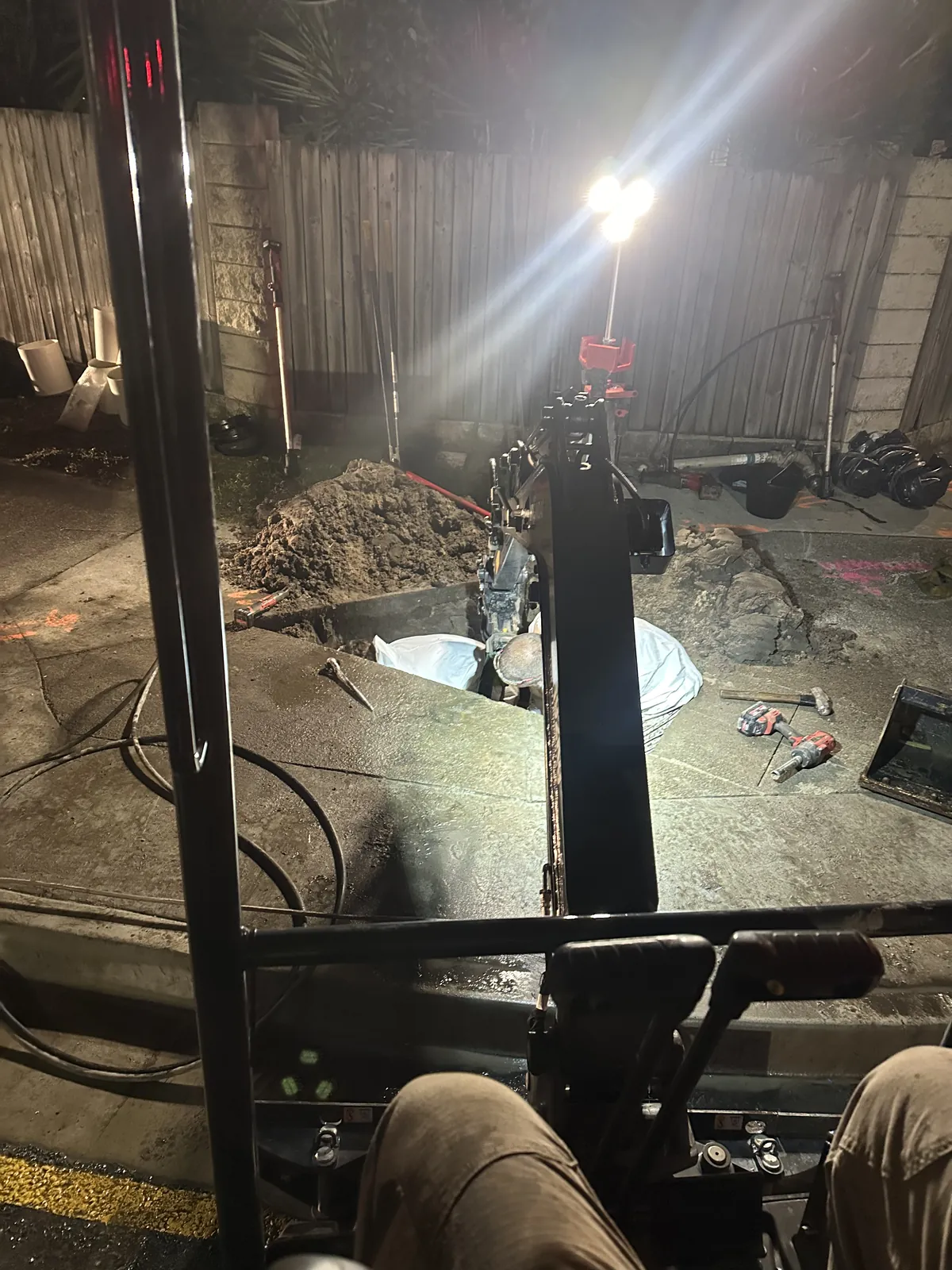 View from a mini excavator cab towards a backyard excavation lit by a lighting tower at night