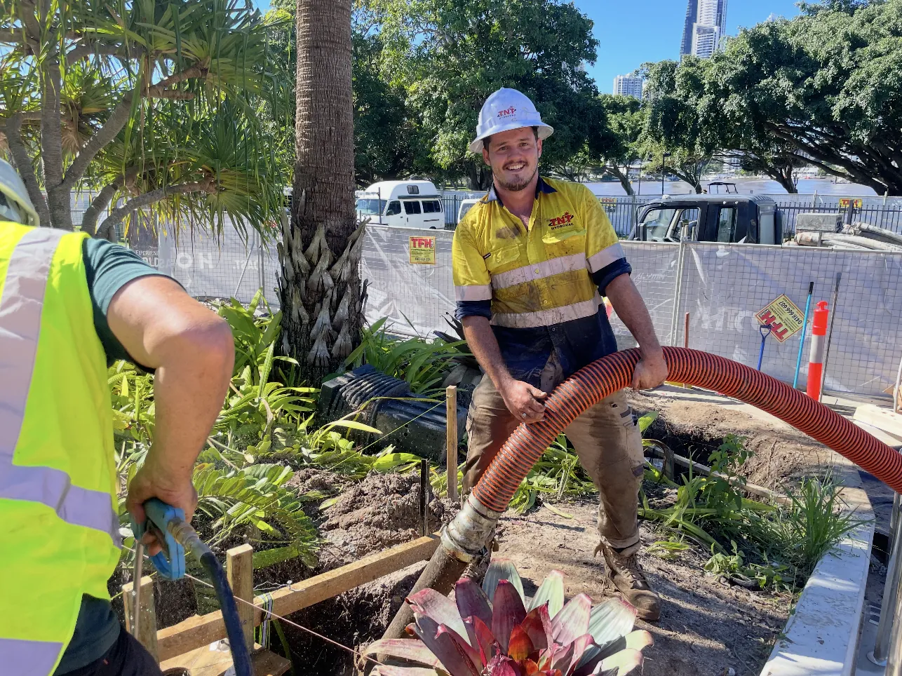 Two workers handling a large orange hose beside excavated ground and a garden bed