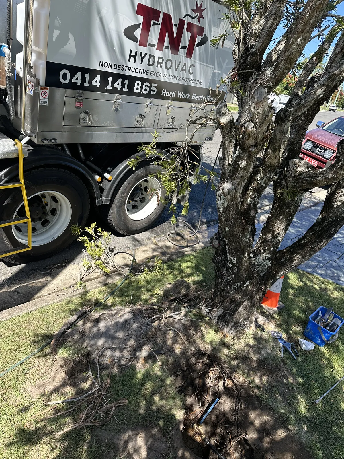 Exposed soil and roots around a tree trunk beside the truck with a hose running into the excavation