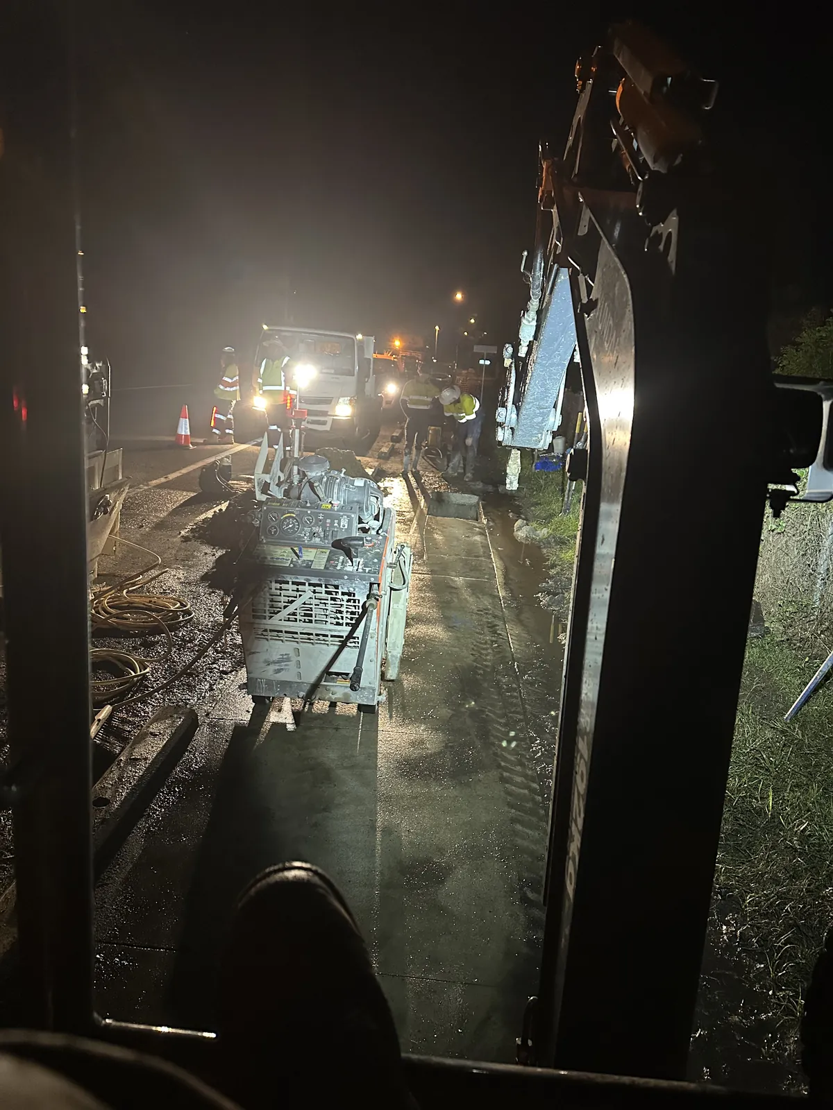 Roadside night works from the cab View from an excavator cab towards lit roadside excavation with workers and equipment at night