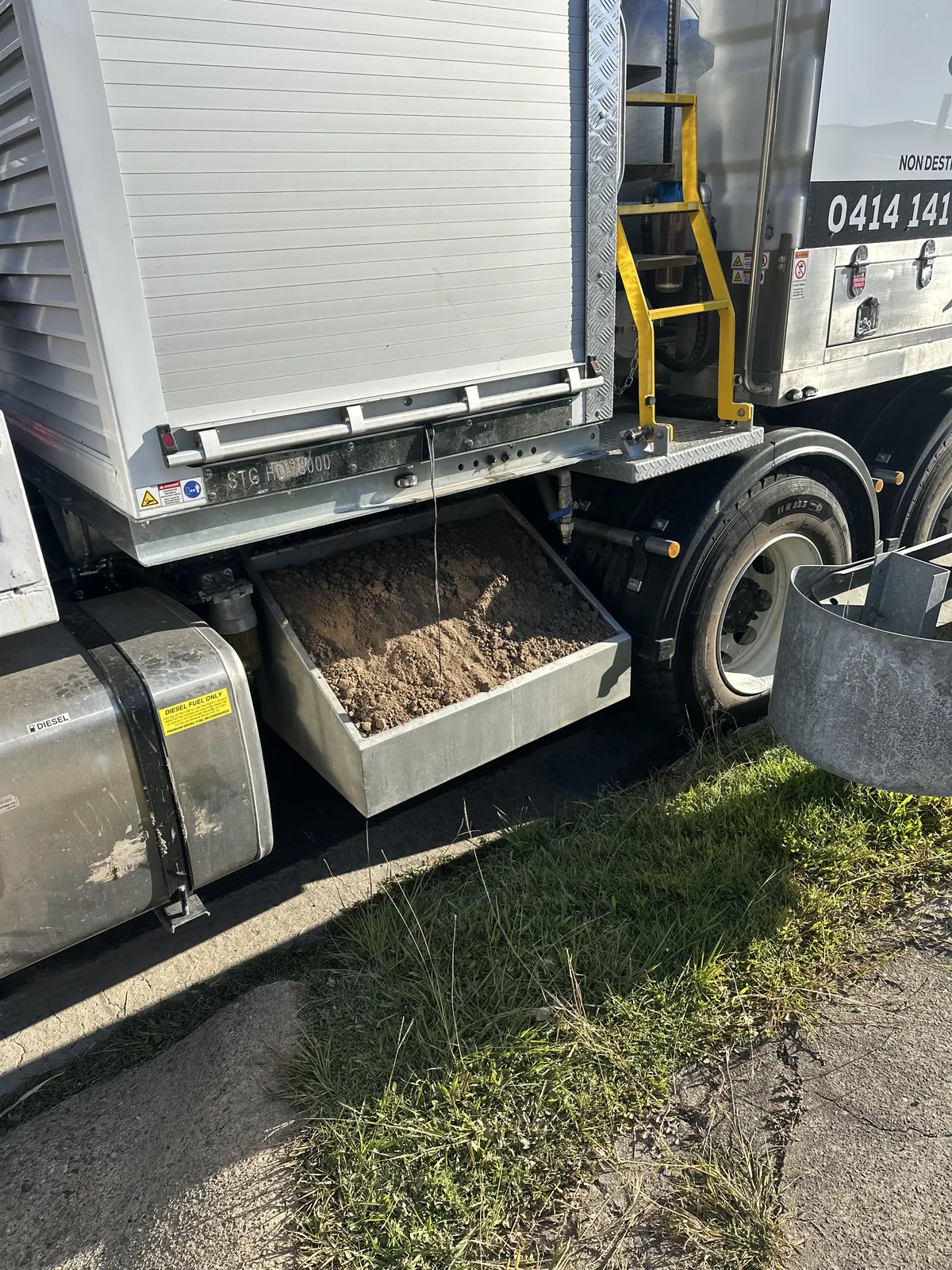 Open metal tray containing sand beneath a truck equipment cabinet, with the ladder and rear wheel beside it