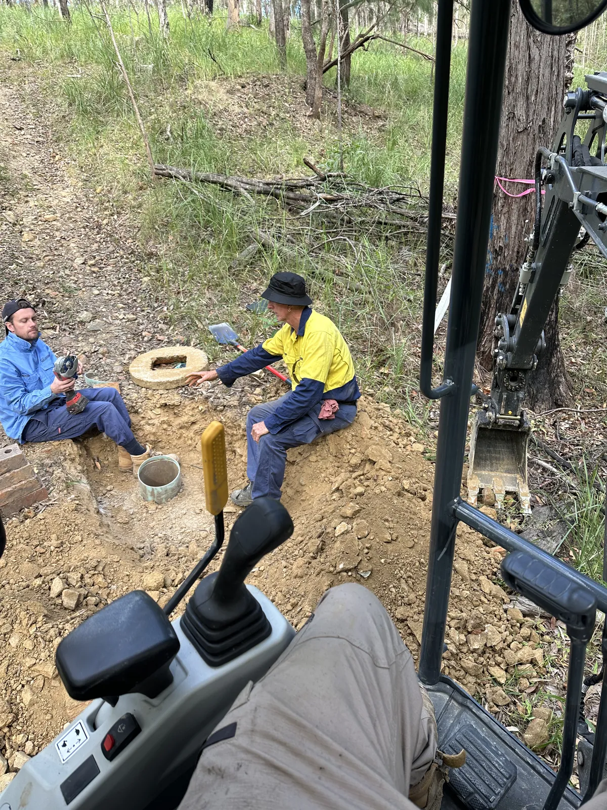 View from an excavator cab towards two workers and an open pit on a wooded track