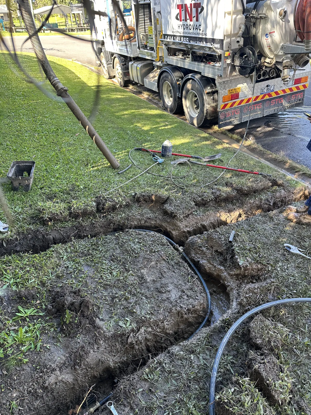Trench with exposed pipes Open trench in grass with dark pipes exposed and hoses leading towards the truck