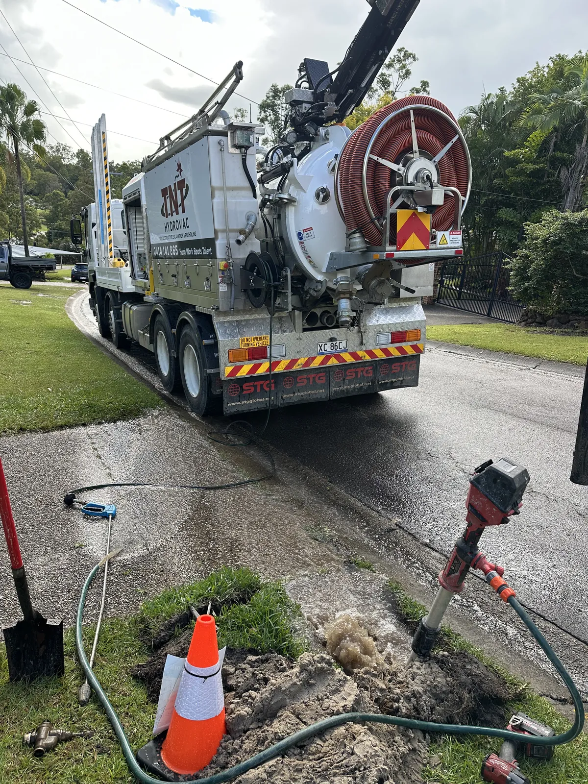 Rear of the truck beside a grass verge with cones, hoses and small ground openings near the kerb