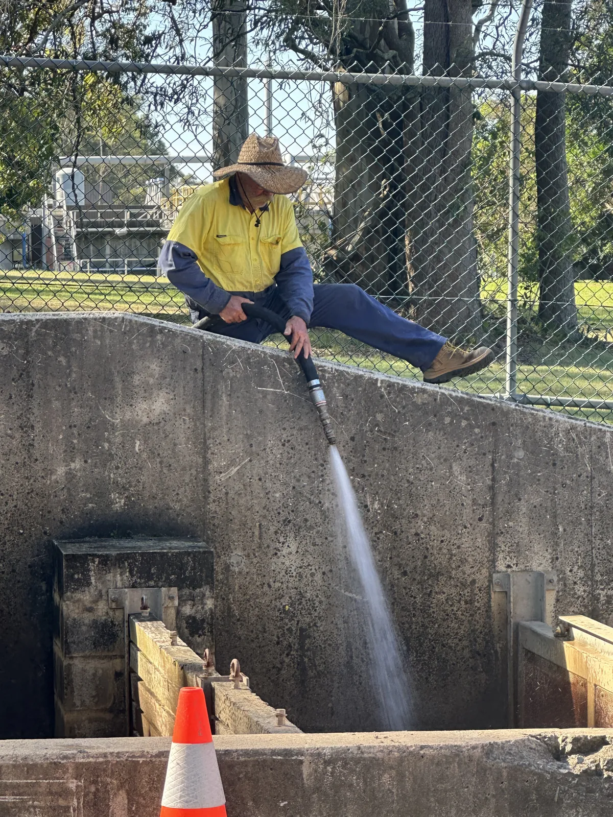 Worker above a concrete structure directing a water lance down into an opening, with a cone below