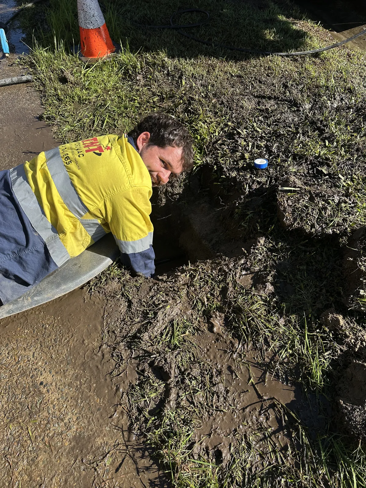Worker in yellow high-visibility clothing leaning beside a muddy ground opening with a hose nearby