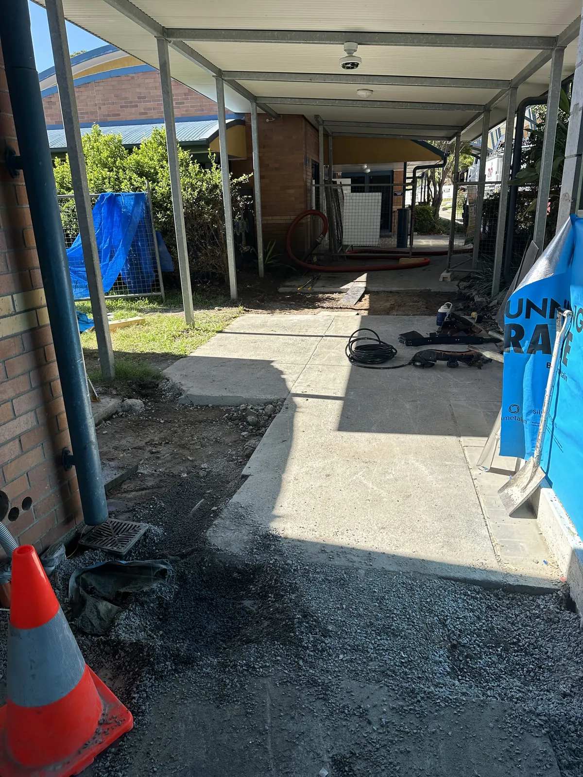 Gravel backfill bordering a concrete path beneath a covered walkway with posts, tools and a traffic cone
