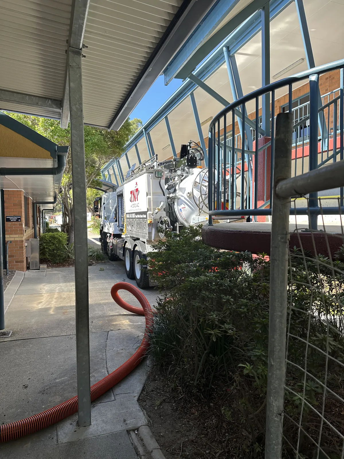 Orange vacuum hose following a covered walkway towards the truck beside a building